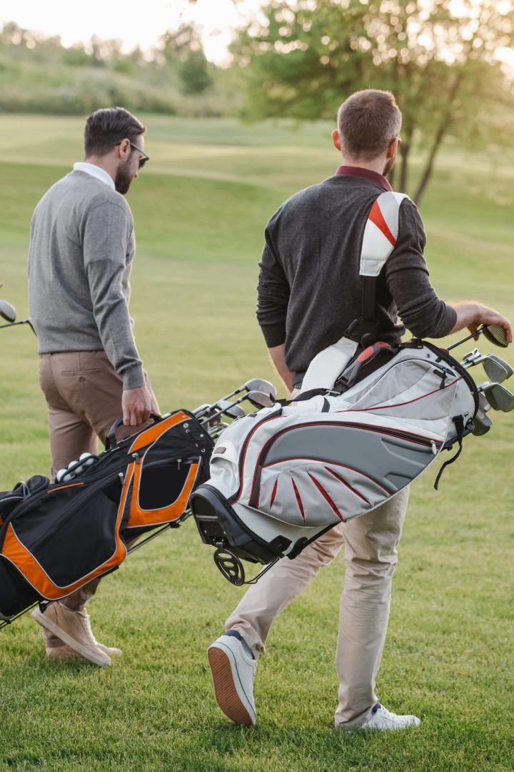 multiethnic golf players with golf clubs in bags walking on golf course